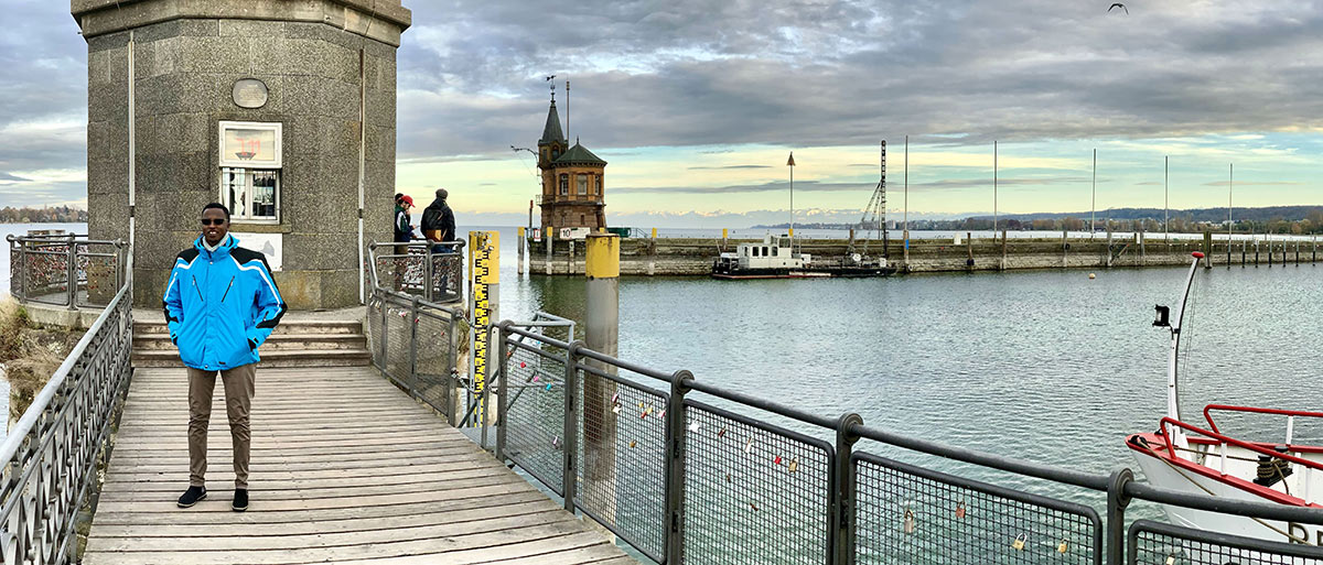 Ole Lerug steht auf einem Steg am Rande des Bodensees, der im Hintergrund zu sehen ist. Ole genießt die kühle Brise am Bodensee.